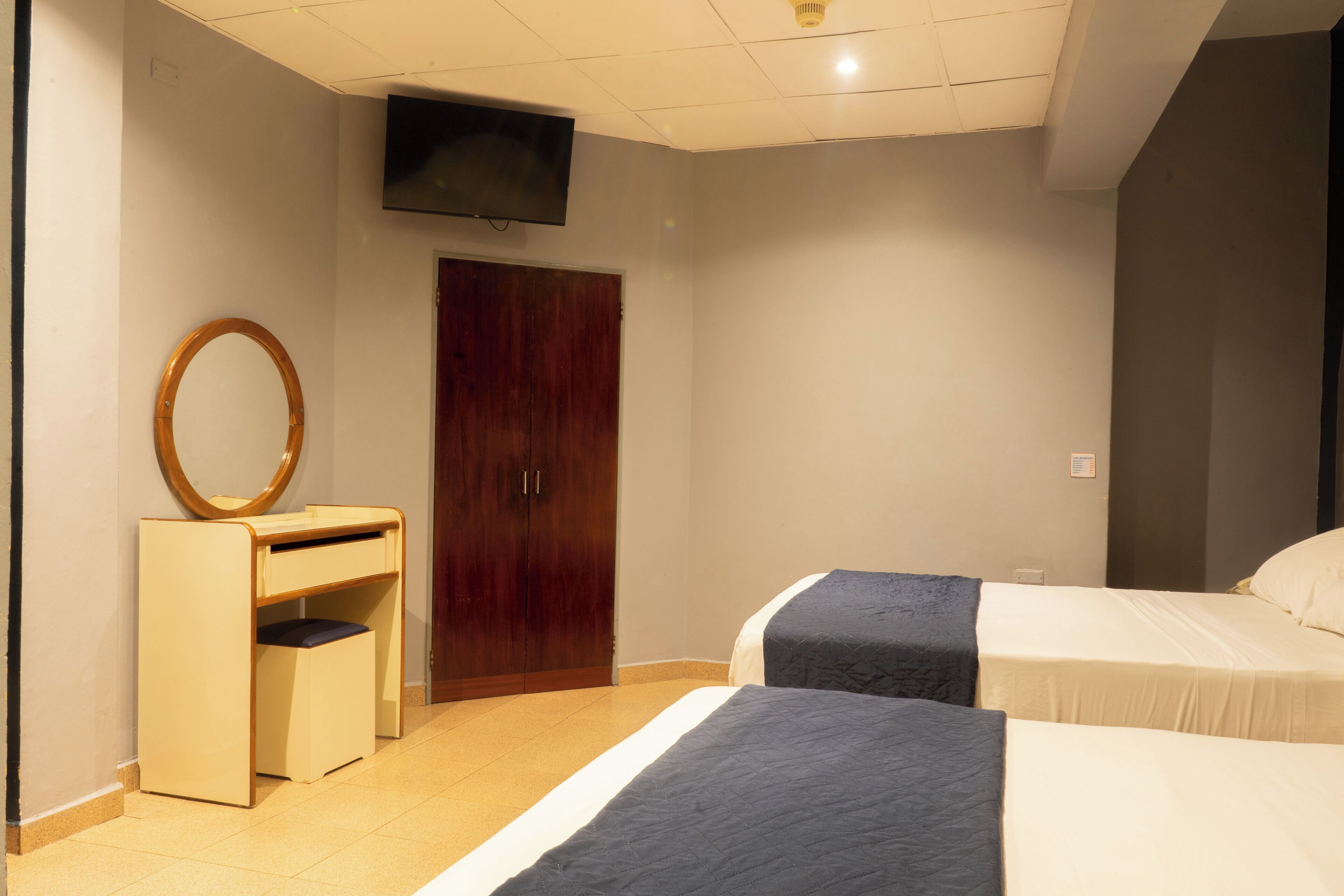 Habitación de hotel con dos camas, un armario de madera, un escritorio pequeño con espejo y un televisor en la pared.