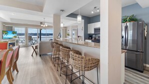 A modern kitchen with a stainless steel refrigerator, a bar area with wicker stools, and a large window overlooking a scenic view. - Harbour Towne 202 - Expansive main channel views! (Lake Ozark)