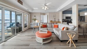 A spacious living area with a large sofa, ottoman, and a small round table. - Harbour Towne 202 - Expansive main channel views! (Lake Ozark)