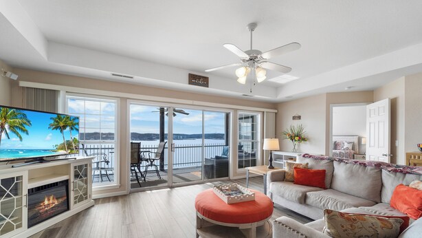 A spacious living room with a large TV, a fireplace, a sofa, and a balcony with a view of the ocean. - Harbour Towne 202 - Expansive main channel views! (Lake Ozark)