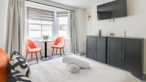 Habitación de hotel con una cama, dos sillones naranjas, una mesita, un televisor y un armario oscuro.