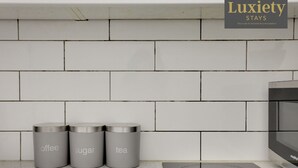 Cocina con salpicadero de azulejos blancos, tres recipientes de acero inoxidable etiquetados con café, azúcar y té, y una moderna cocina de inducción.