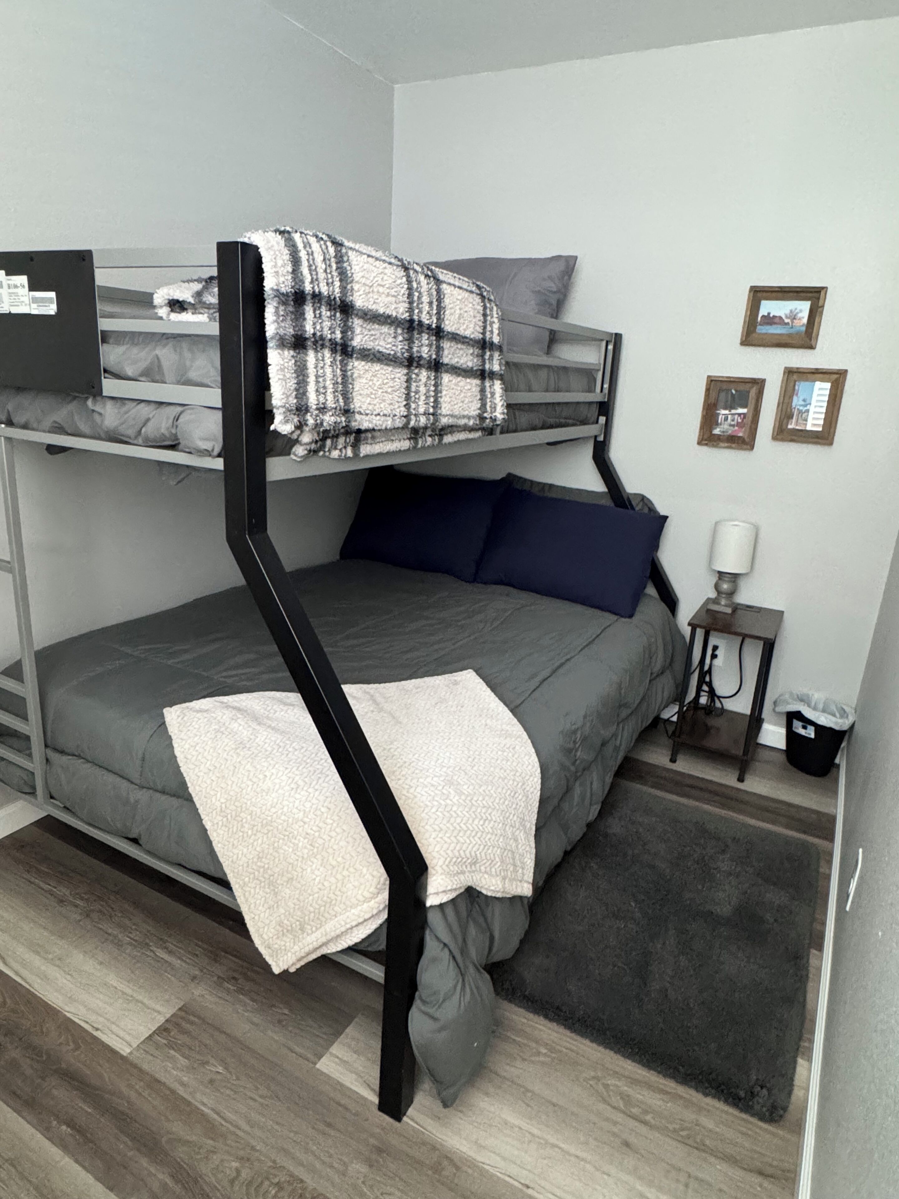 A bunk bed with a grey mattress, a white throw blanket, and a plaid blanket on top. There is a small bedside table with a lamp and a trash bin.