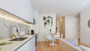 Una cocina moderna con armarios blancos, encimera de mármol y horno integrado. Hay un comedor con mesa y sillas blancas, y un sofá en la sala de estar.