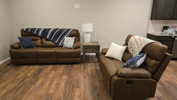 A living room with two brown leather sofas, a small wooden side table with a lamp, and a kitchen area in the background. - The Benjamin House, Quality and Comfortable Townhouse with small town feel (Hallsville)