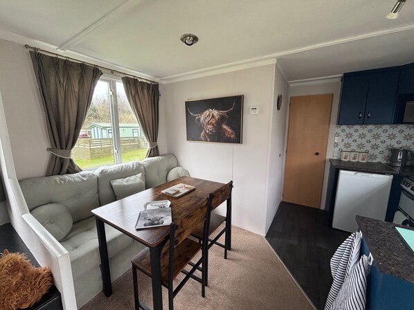 A compact living space with a sofa, a dining table, a kitchen area, and a window with curtains. - No 38 Heilan Hoose at Three Lochs Newton Stewart (Newton Stewart)