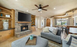 A living room with a stone fireplace, a flat-screen TV, a ceiling fan, and a dining area with a bar.