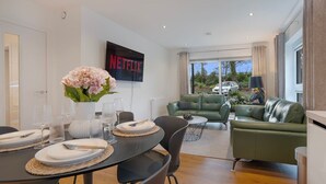 A modern living room with a dining area, green leather sofas, a flat-screen TV, and a large window with a view.