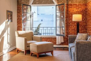 A room with a brick wall, a window with open shutters, a beige armchair, a grey sofa, and a floor lamp.