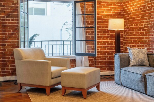 A room with a brick wall, a large window with a view, a beige armchair, a grey sofa, and a floor lamp.