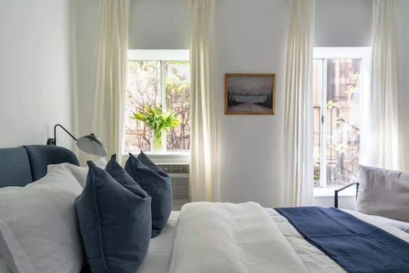 A bedroom with a bed, pillows, a bedside lamp, a window with curtains, and a painting on the wall.