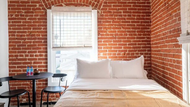 Une petite pièce agréable, avec un mur en briques, un lit simple recouvert de linge de lit blanc, une petite table avec deux chaises et une fenêtre ornée d’une grille métallique.