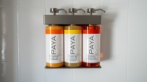 Trois bouteilles de revitalisant, de gel douche et de shampoing de la marque Paya sont posées sur une étagère, devant un mur recouvert de carreaux blancs.
