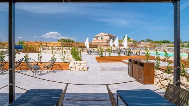 A covered outdoor seating area with lounge chairs and umbrellas, a stone wall, and a view of a building and pool in the background.