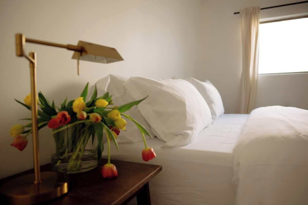 A hotel room with a bed, white bedding, a bedside table with a lamp, and a vase with flowers.