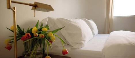 A hotel room with a bed, white bedding, a bedside table with a lamp, and a vase with flowers.