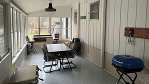 Une pièce avec une table et des chaises, un ventilateur de plafond, un barbecue et une fenêtre donnant sur des arbres.