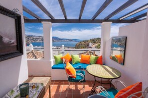 Ein Balkon mit einer weißen Wand, einer blauen gepolsterten Bank mit orangefarbenen und grünen Kissen, einem runden Tisch mit einer grün gemusterten Tischdecke und Blick auf das Meer und die Hügel.