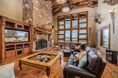 A rustic interior with a stone fireplace, wooden entertainment unit, leather sofa, and large windows offering a view.. The Wildflower, Lodge+guest cabins 26 people