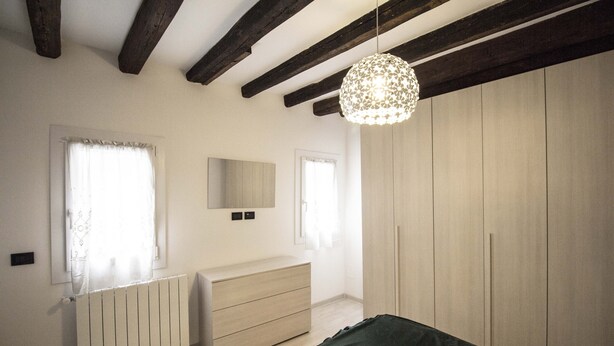 Ein Schlafzimmer mit Holzbalkendecke, Heizkörper, Kommode, Bett und Kleiderschrank.