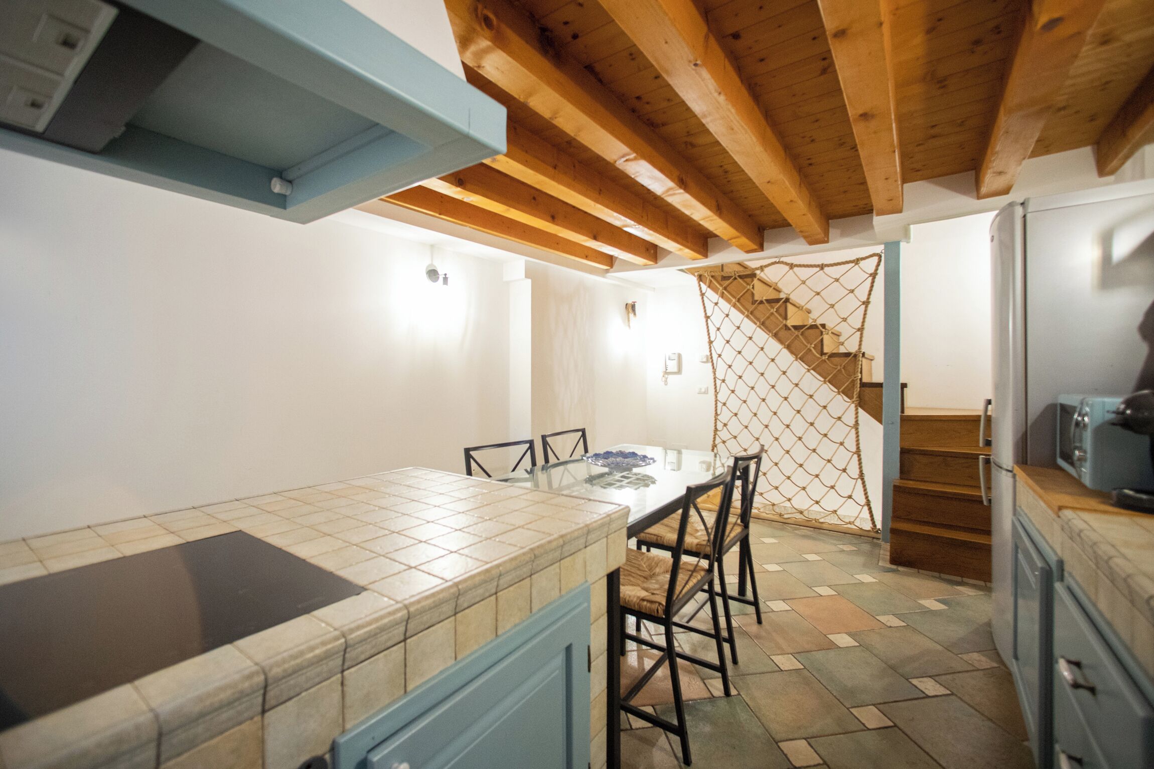 Cucina con piano di lavoro in piastrelle, travi in legno a soffitto, tavolo da pranzo con sedie e una scala che conduce ad un altro livello.