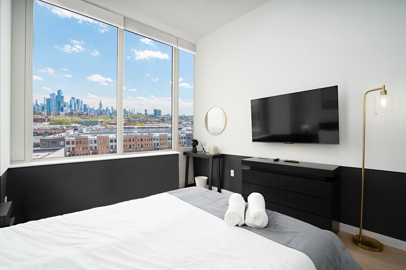 Habitación de hotel moderna con una cama grande, un televisor montado en la pared y vistas a la silueta urbana a través de amplios ventanales.
