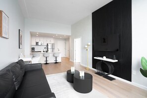 A modern living room with a black sofa, a coffee table, and a kitchen in the background. - Exclusive 1BR Apt | Midtown NYC Skyline View (Hoboken)