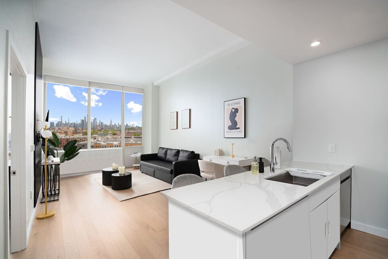 A modern apartment with a kitchen, dining area, and living room. It features a large window with a city view, a black leather sofa, and a white island with a sink.