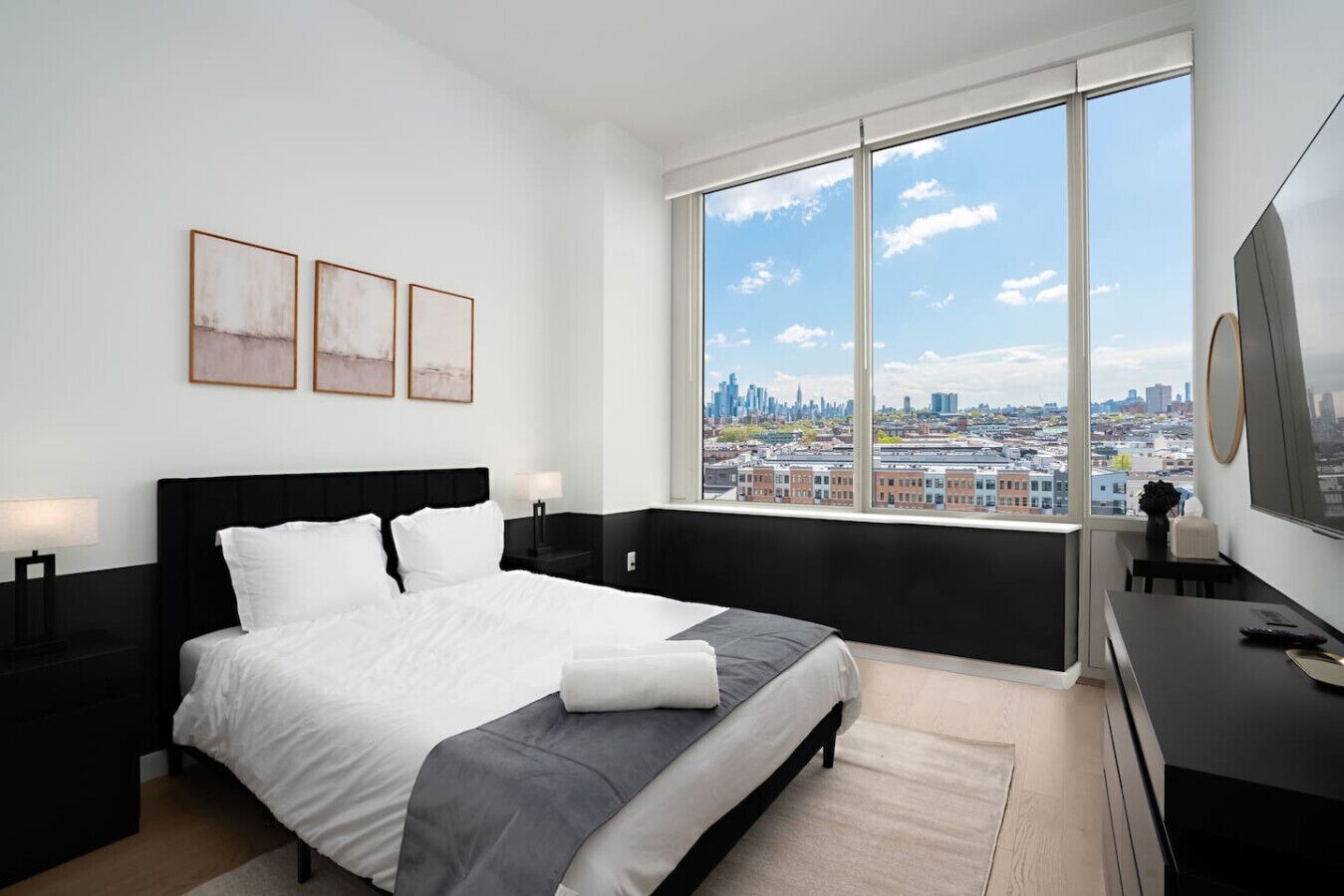 A modern bedroom with a large bed, a view of the cityscape through a floor-to-ceiling window, and a flat-screen TV on a dresser.