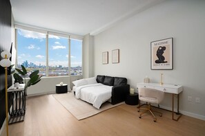 A modern bedroom with a large window, a black leather sofa, a white desk, and a framed artwork on the wall.