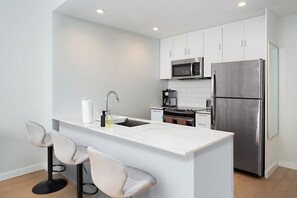 A modern kitchen with white cabinets, a stainless steel refrigerator, a microwave, and an oven. There are two bar stools at the island.