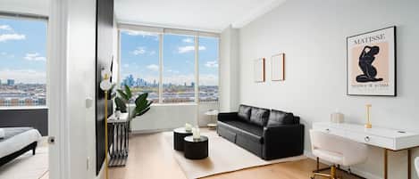 A modern living room with a black leather sofa, white desk, and a large window offering a city view.