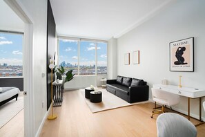 A modern living room with a black leather sofa, white desk, and a large window offering a city view. - Exclusive 1BR Apt | Midtown NYC Skyline View (Hoboken)