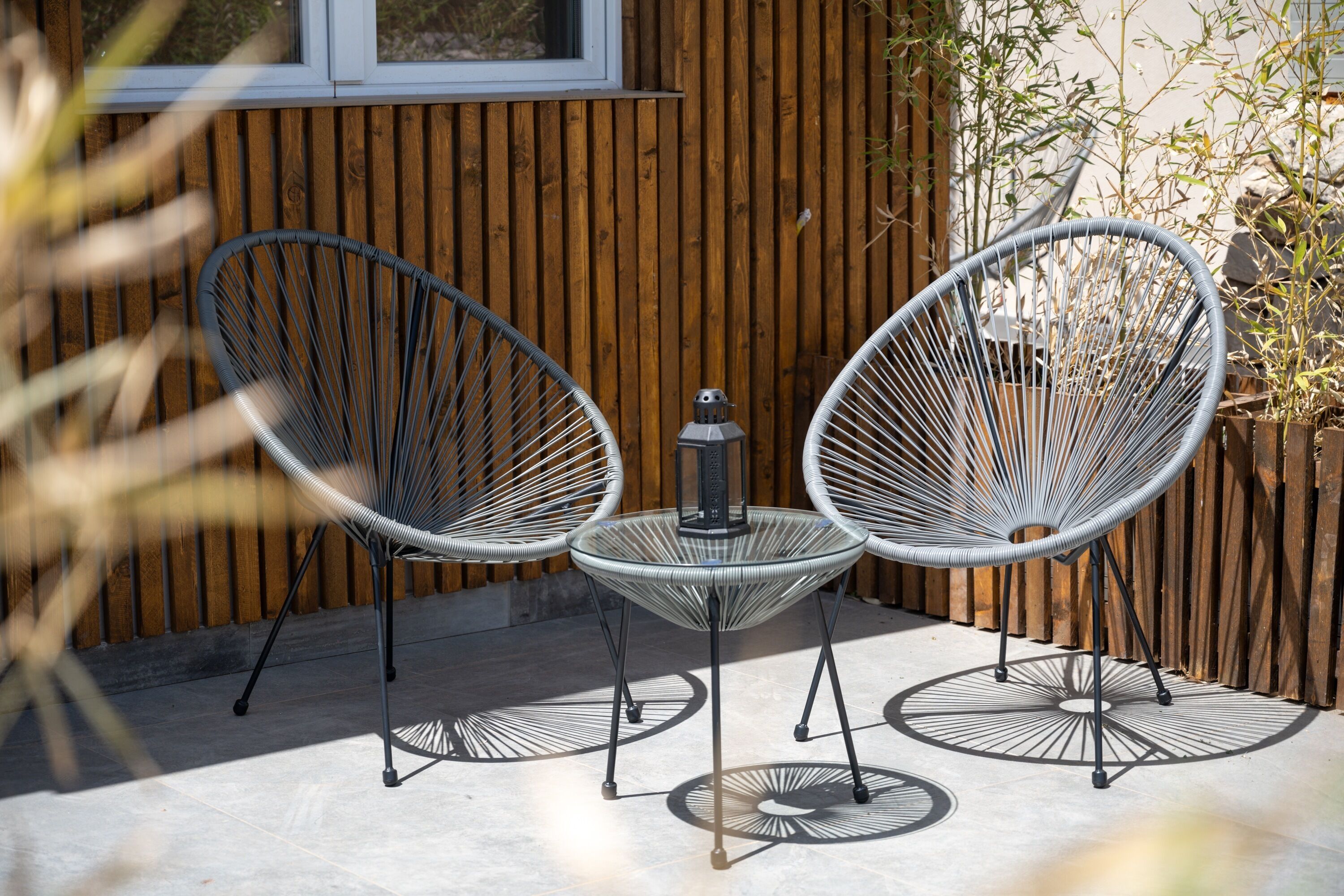 A patio with two wire-framed chairs and a small table, set against a wooden wall with a window above.