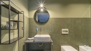 Et badeværelse med en hvid vask, et toilet og et spejl. Der er en hylde med genstande på, og en lampe over spejlet.