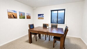 Uma sala de jantar com uma mesa retangular e quatro cadeiras, chão coberto por carpete e quatro pinturas de paisagens nas paredes.