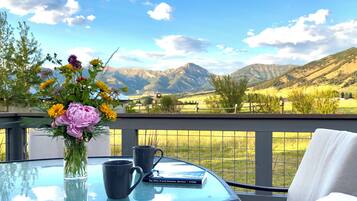 Een glazen tafel met een bloevas, twee mokken en een boek getiteld 'Day Hikes Around Bozeman, Montana' op een balkon met uitzicht op bergen.