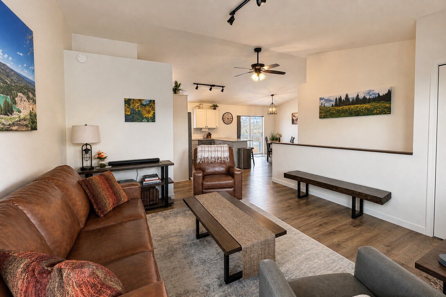 A living room with a brown leather sofa, a wooden coffee table, and a bench. There is a kitchen in the background with a dining area.