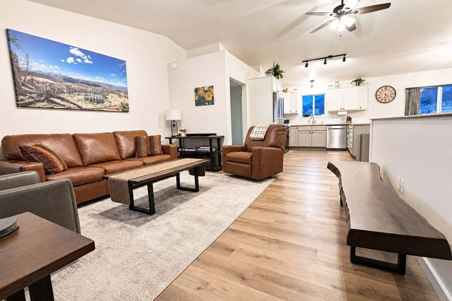 Un salon moderne avec un canapé en cuir, une table basse et un meuble pour la télévision. On aperçoit une cuisine à l’arrière-plan, avec des meubles blancs et une horloge au mur.