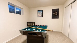 Uma mesa de bilhar em um ambiente com piso coberto por carpete, uma janela e duas quadros pendurados nas paredes.