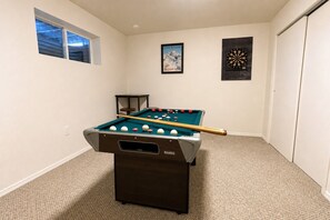 Une table de billard dans une pièce dont le sol est recouvert de moquette, avec une fenêtre et deux tableaux encadrés accrochés aux murs.