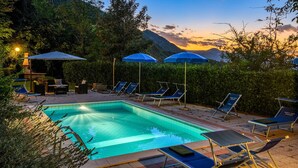 A pool area with lounge chairs and umbrellas, surrounded by greenery and a view of mountains at sunset. - Acquanera 6&2 by Marche Holiday Villas (Piobbico)