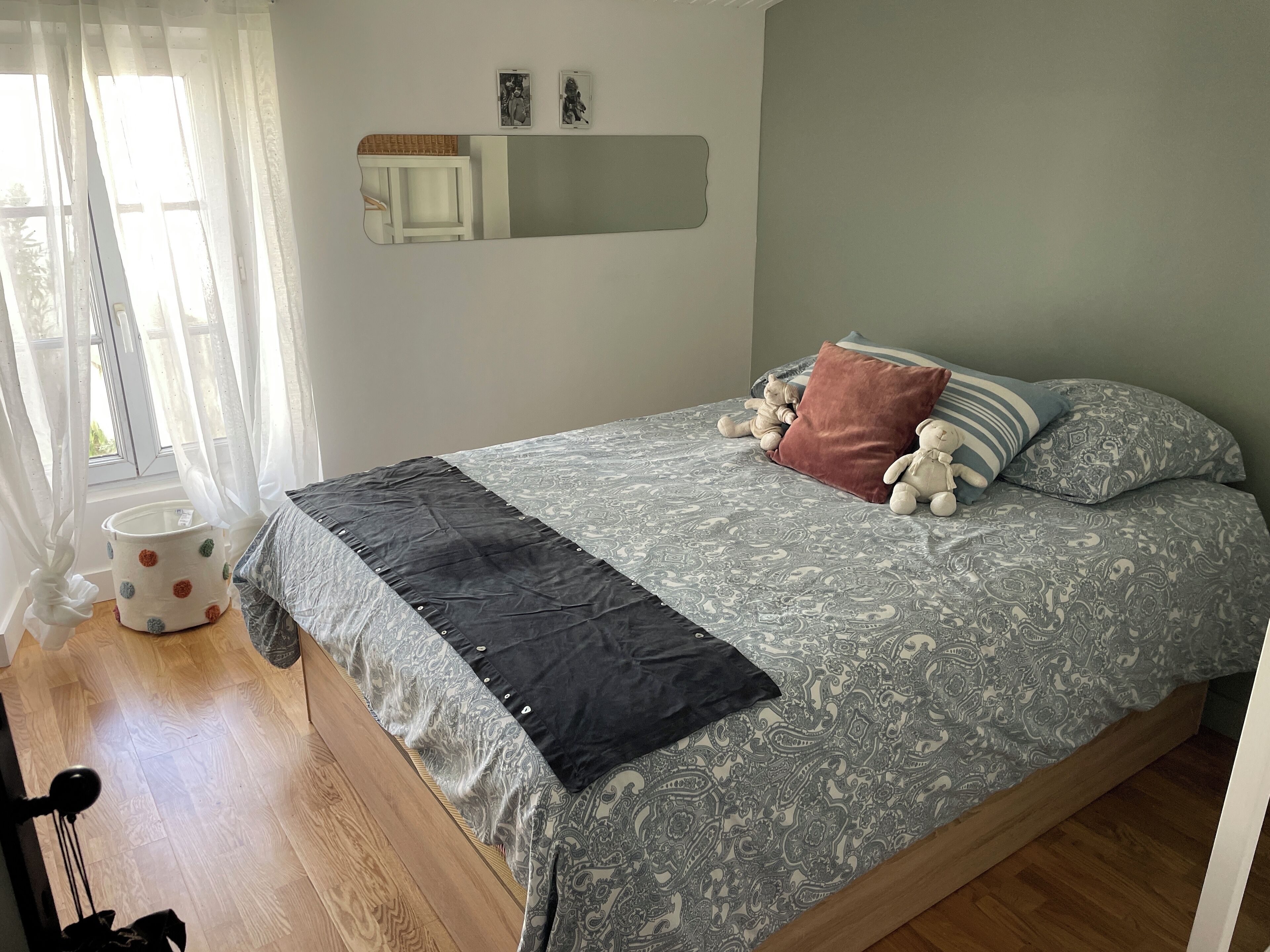 A bedroom with a bed, pillows, a bedside table, a mirror, and a window with curtains.