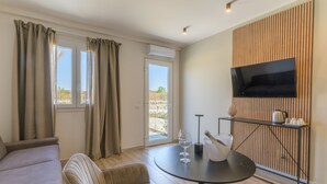 En moderne stue med sofa, lænestole, spisebord med vinglas, et tv monteret på en væg af træpaneler, og en dør der fører til et udendørs område.