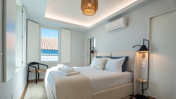A modern bedroom with a large bed, a chair, an air conditioning unit, and a window with a view of a building.