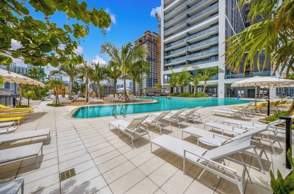 A rooftop pool area with multiple lounge chairs and umbrellas, surrounded by palm trees and modern buildings. - Luxe 1-BR with Stunning Downtown Views (Miami)