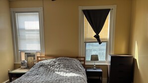 A bedroom with a bed, bedside table, lamp, dresser, and windows with blinds.
