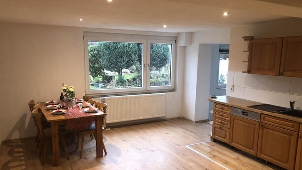 Un comedor con una mesa y sillas de madera, una ventana con vista a follaje, y una cocina con gabinetes de madera.