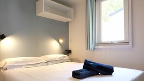 Een keurig opgemaakt bed met witte lakens, een blauwe handdoek gevouwen en een wandplank erboven.