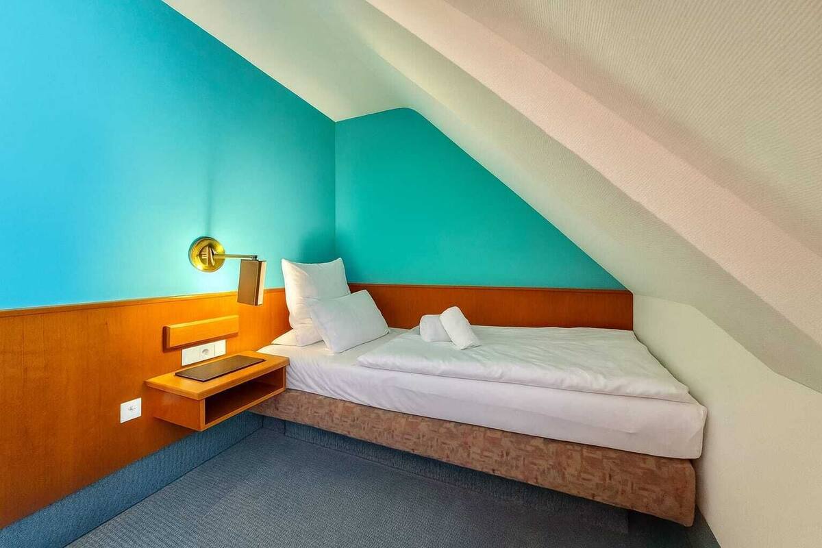 A small, cozy room with a single bed, a nightstand, and a wall-mounted lamp.